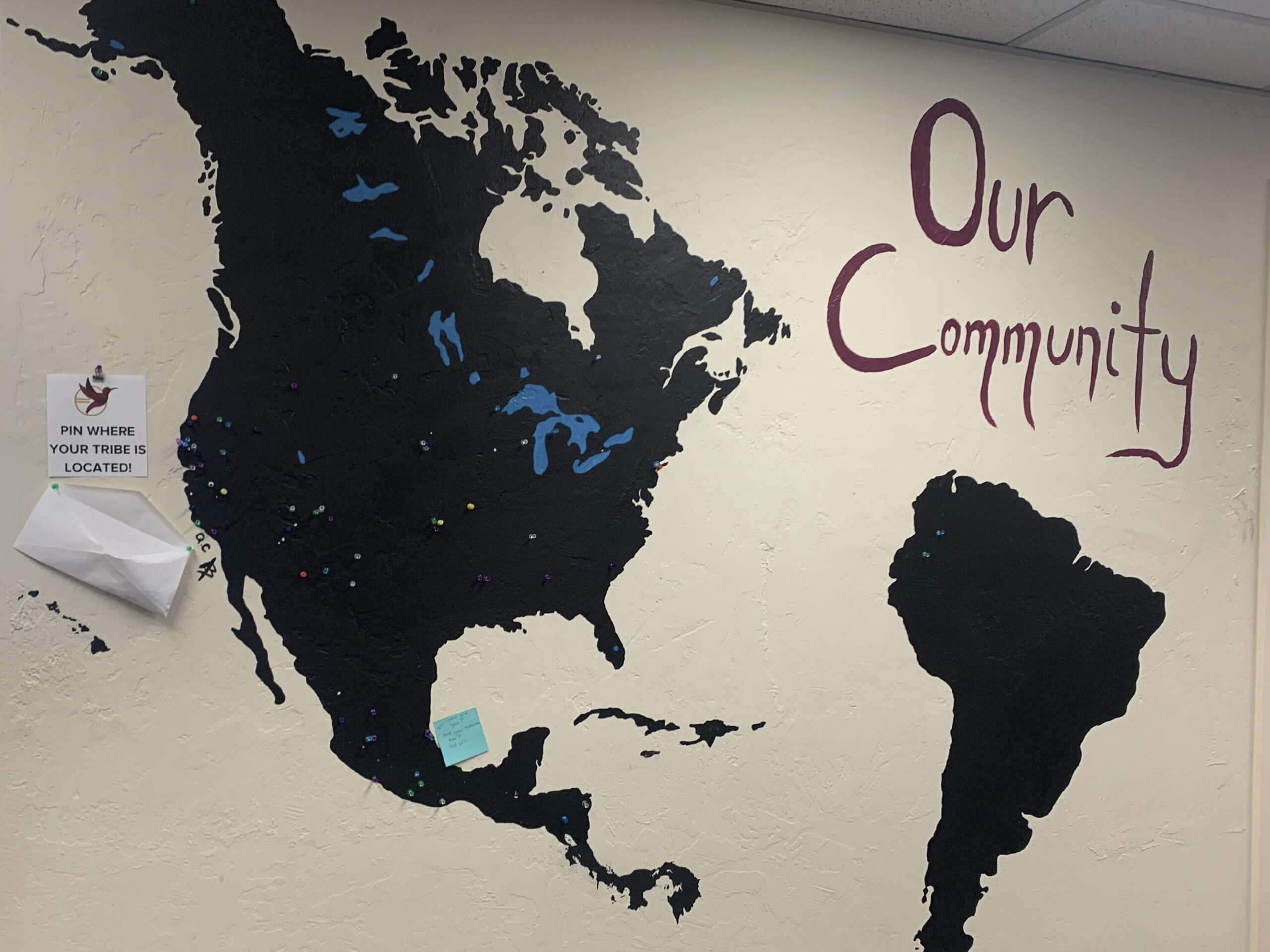 A mural of North and South America painted on a white wall with the words “our community” above it. Bright blue push pins are placed on the regions where each student’s people are from.