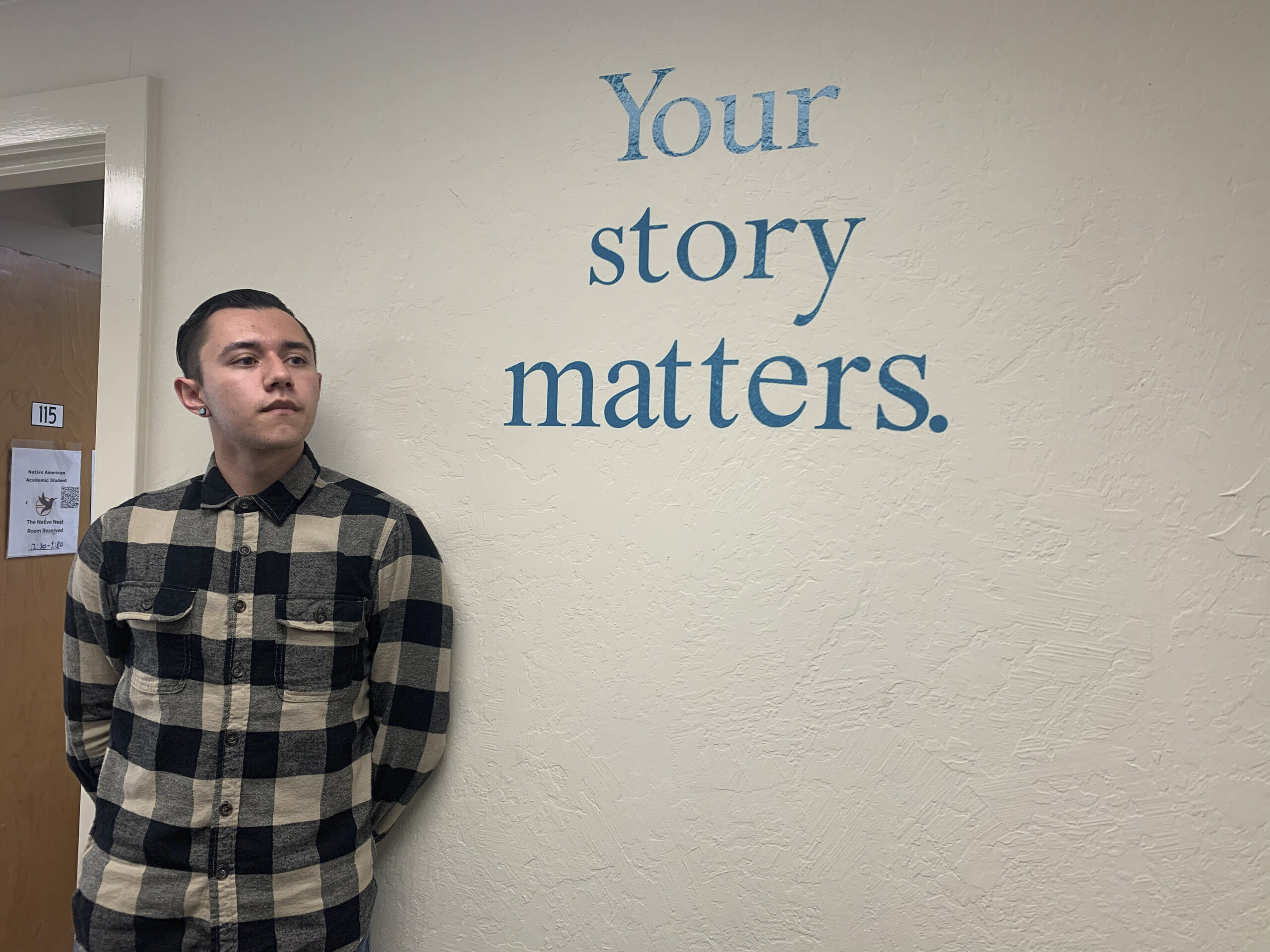 A young Cherokee man standing in front of the phrase “your story matters” that is painted on a white wall.