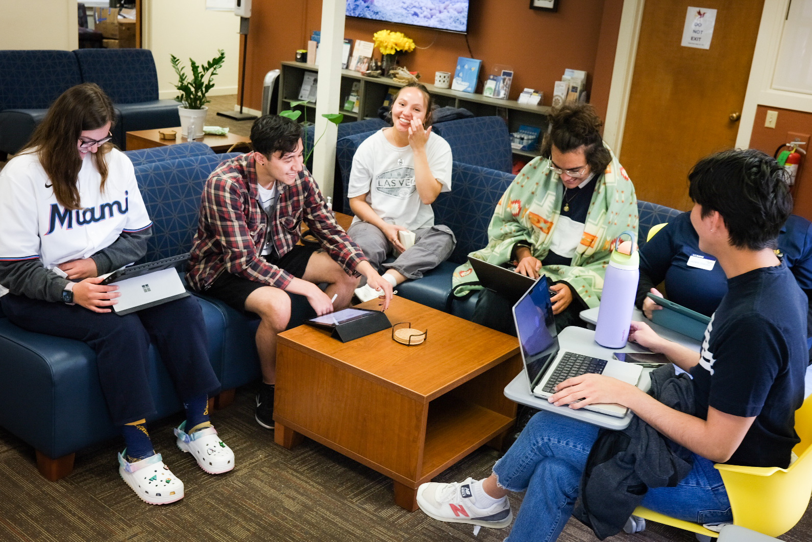 A group of Native students sitting on blue couches in the Native Nest’s lounge. The students are talking and laughing as they work on their computers and tablets.